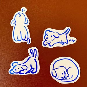 以下が含まれることがあります： 白い紙のステッカーに、青い線で描かれた犬のイラストが4つあります。犬はそれぞれ異なるポーズをとっています。1匹は後ろ足で立ち、1匹は走っています。1匹は横たわっていて、1匹は丸まっています。