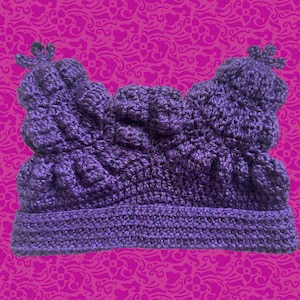 May include: A handmade, dark purple crochet hat with a textured design. The hat features a wide brim and decorative elements on top. The background is a patterned pink surface.