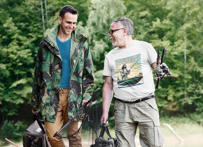 May include: Two people walking in a forest, one wearing a camouflage jacket and the other wearing a white t-shirt with the text "Slingin' Poles Catchin' Souls".