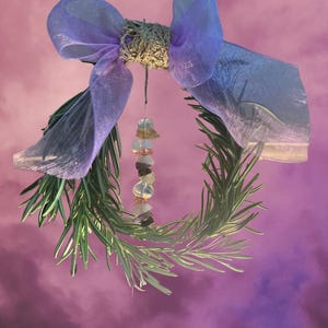 May include: A green rosemary wreath with a purple and blue sheer ribbon bow and a crystal charm hanging in the center. The wreath is against a pink background.