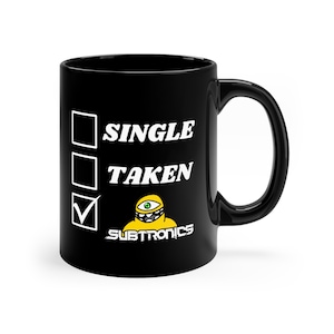 May include: Black ceramic mug with a white check box and the text "SINGLE" and "TAKEN". The check box next to "TAKEN" is checked. A yellow cartoon character with a single eye and a toothy grin is below the check box. The text "SUBTRONICS" is below the cartoon character.