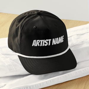 May include: Black baseball cap with white embroidered text that reads "ARTIST NAME". The cap has a white rope detail along the brim and a curved visor. The hat is sitting on a white surface.