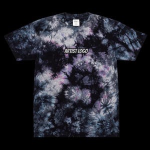 May include: A tie-dye t-shirt in a pattern of black, white, and purple. The short-sleeved shirt has a crew neck. The text "ARTIST LOGO" is printed across the chest in a white, block font.