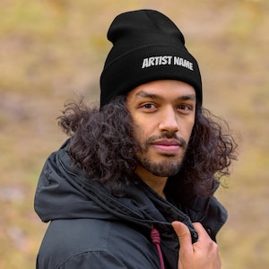 May include: Black beanie with the text "ARTIST NAME" in white. The beanie is worn by a person with long, curly hair and a black jacket. The background is blurred, with a natural, outdoor setting.
