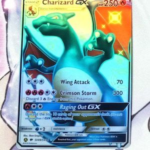May include: A holographic Pokemon trading card featuring Charizard GX, a large, blue, fire-breathing dragon with orange wings. The card has a gold border and features the text "Charizard GX" and "HP 250". The card also has three attacks: "Wing Attack", "Crimson Storm", and "Raging Out GX".