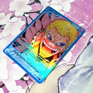 May include: A blue and pink anime trading card featuring Donquixote Doflamingo from the One Piece series. The card has a special attack power of 5000 and a life value of 0. The card text reads "DON!! x2 When Attacking: 1) You may reveal the top card of your deck if that card is a (The Seven Warlords of the Sea) type Character card with a cost of 4 or less, you may play that card rested."