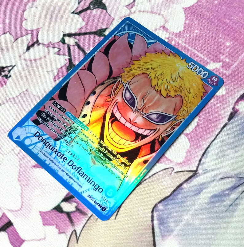 May include: A blue and pink anime trading card featuring Donquixote Doflamingo from the One Piece series. The card has a special attack power of 5000 and a life value of 0. The card text reads "DON!! x2 When Attacking: 1) You may reveal the top card of your deck if that card is a (The Seven Warlords of the Sea) type Character card with a cost of 4 or less, you may play that card rested."