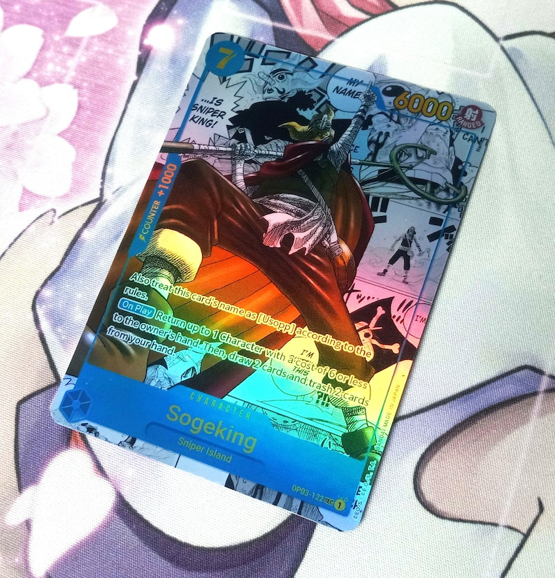 Pode incluir: Um cart&atilde;o de negocia&ccedil;&atilde;o hologr&aacute;fico com um personagem do anime One Piece. O cart&atilde;o &eacute; azul e apresenta um personagem chamado Sogeking, que &eacute; um atirador de elite. O cart&atilde;o tem um n&iacute;vel de poder de 6000 e um valor de contador de +1000. O texto do cart&atilde;o diz: "Tamb&eacute;m trate o nome deste cart&atilde;o como [Usopp] de acordo com as regras. Ao jogar, devolva at&eacute; 1 personagem com um custo de 6 ou menos para a m&atilde;o do propriet&aacute;rio. Em seguida, compre 2 cartas e descarte 2 cartas de sua m&atilde;o."