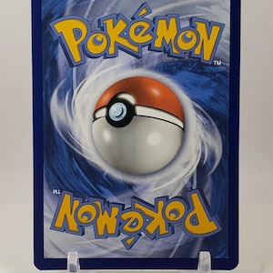 Puede incluir: El reverso de una carta coleccionable de Pok&eacute;mon, con un borde azul y un fondo blanco y azul en espiral. El logotipo amarillo de Pok&eacute;mon y una Pok&eacute; Ball roja y blanca est&aacute;n en el centro. La tarjeta se muestra en un soporte de pl&aacute;stico transparente.