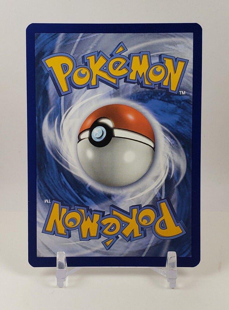 Puede incluir: El reverso de una carta coleccionable de Pok&eacute;mon, con un borde azul y un fondo blanco y azul en espiral. El logotipo amarillo de Pok&eacute;mon y una Pok&eacute; Ball roja y blanca est&aacute;n en el centro. La tarjeta se muestra en un soporte de pl&aacute;stico transparente.