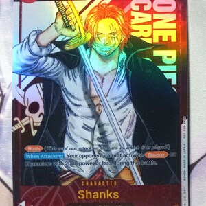 May include: A collectible trading card featuring Shanks from the anime One Piece. The card is red, white, and blue with a holographic finish. The card features a picture of Shanks with his signature red hair and a sword. The card is numbered 000/000 and has a power level of 10000. The card text reads "Rush (This card can attack on the turn in which it is played.) When Attacking Your opponent cannot activate Blocker on characters with 2000 power or less during this battle."