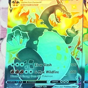May include: A holographic Charizard VMAX Pokémon card. The card features a fiery dragon illustration with the text "Charizard VMAX" and "Claw Slash" and "G-Max Wildfire" attack descriptions. The card has a HP of 330.