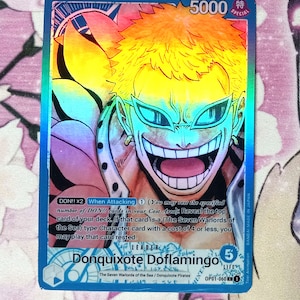 May include: A blue and pink anime trading card featuring Donquixote Doflamingo from the One Piece series. The card has a holographic finish and features the text "DON!! x2 When Attacking 1 (You may rest the specified number of DON!! cards in your Cost Area): Reveal the top card of your deck. If that card is a (The Seven Warlords of the Sea) type Character card with a cost of 4 or less, you may play that card rested."  The card also features the text "LEADER Donquixote Doflamingo 5 LIFE OP01-060 1".