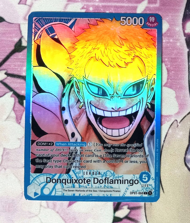 May include: A blue and pink anime trading card featuring Donquixote Doflamingo from the One Piece series. The card has a holographic finish and features the text "DON!! x2 When Attacking 1 (You may rest the specified number of DON!! cards in your Cost Area): Reveal the top card of your deck. If that card is a (The Seven Warlords of the Sea) type Character card with a cost of 4 or less, you may play that card rested."  The card also features the text "LEADER Donquixote Doflamingo 5 LIFE OP01-060 1".