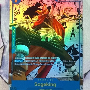 Pode incluir: Um cart&atilde;o de troca com uma ilustra&ccedil;&atilde;o colorida de Sogeking, um personagem do anime One Piece. O cart&atilde;o &eacute; azul e branco com o texto "Sogeking" e "Sniper Island". O cart&atilde;o tem um n&iacute;vel de poder de 6000 e um valor de contador de +1000.