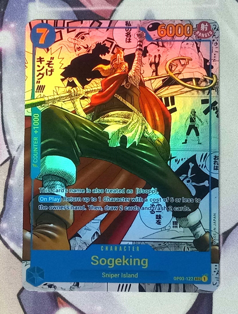 Pode incluir: Um cart&atilde;o de troca com uma ilustra&ccedil;&atilde;o colorida de Sogeking, um personagem do anime One Piece. O cart&atilde;o &eacute; azul e branco com o texto "Sogeking" e "Sniper Island". O cart&atilde;o tem um n&iacute;vel de poder de 6000 e um valor de contador de +1000.