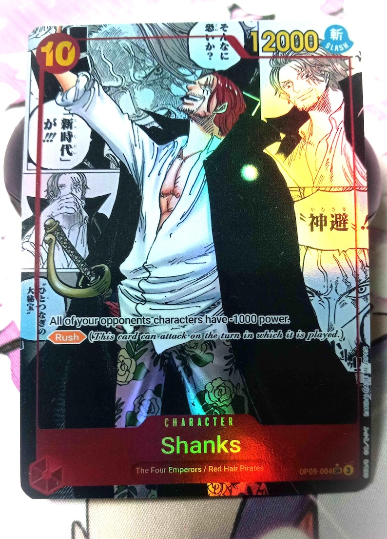 Puede incluir: Una carta de intercambio roja, blanca y negra con un personaje del anime One Piece. El personaje es Shanks, un pirata pelirrojo con un abrigo negro y una camisa blanca. La carta tiene un acabado hologr&aacute;fico y el texto "Shanks" en letras rojas. La carta tambi&eacute;n tiene el texto "The Four Emperors / Red Hair Pirates" y "OP09-004".