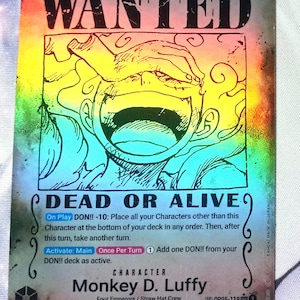 May include: Holographic trading card featuring Monkey D. Luffy from the anime series One Piece. The card reads "WANTED" with the text "DEAD OR ALIVE" below a large illustration of Luffy. The card has a value of 10 and 12000 strike.