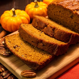 May include: A wooden cutting board with slices of pumpkin bread, two whole mini pumpkins, and pecans scattered around the board.