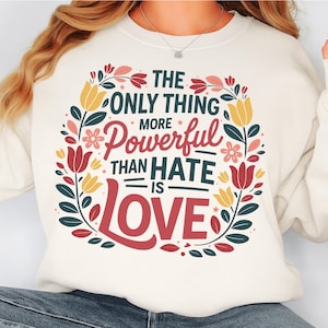 May include: Cream-coloured sweatshirt featuring a floral wreath design. The wreath frames the text "The Only Thing More Powerful Than Hate Is Love" in red, green, and white. The sweatshirt is a casual style.
