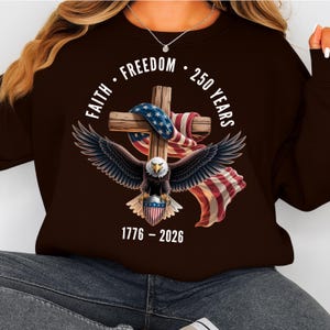 May include: A brown sweatshirt featuring a patriotic design. The graphic includes a wooden cross draped with an American flag, an eagle with outstretched wings, and the text "Faith Freedom 250 Years 1776-2026."