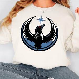 May include: Cream-colored sweatshirt featuring a graphic of a black bird with outstretched wings, a red eye, and a blue star above it. The bird is set within a blue and black crescent design. The sweatshirt is a casual style.