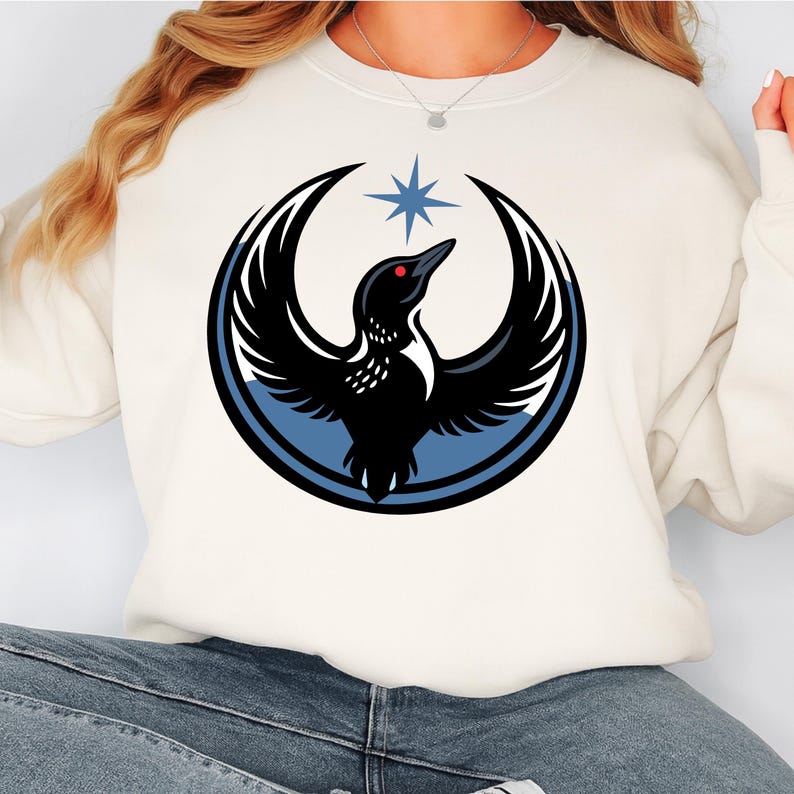 May include: Cream-colored sweatshirt featuring a graphic of a black bird with outstretched wings, a red eye, and a blue star above it. The bird is set within a blue and black crescent design. The sweatshirt is a casual style.