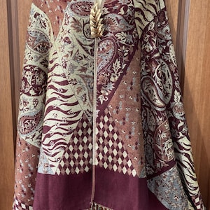 May include: A rectangular shawl featuring a patchwork design of burgundy, tan, and cream colours. The fabric displays paisley, diamond, and animal print patterns with a gold-toned wheat-shaped brooch at the centre and fringe along the bottom edge.