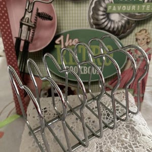 May include: A chrome toast rack with six slots, positioned in front of a vintage cookbook-themed background. The rack is on a white lace doily, with the words "Women's Weekly" visible.