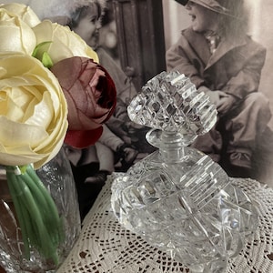 May include: A clear, cut-glass perfume bottle with a diamond pattern. The bottle has a decorative stopper and sits on a white lace doily. A vase of flowers is to the left, and a vintage black and white photo is in the background.