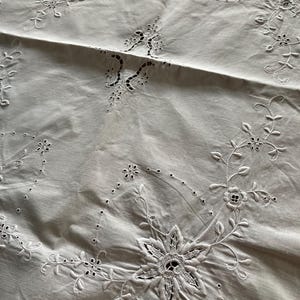 May include: White embroidered tablecloth with floral and leaf motifs. The design incorporates large, openwork flowers and delicate vines with small blossoms. The cloth has a soft, slightly wrinkled texture, suggesting a vintage or handmade quality.