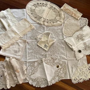 May include: A collection of white and off-white linens including embroidered napkins, lace doilies, and table runners. The pieces feature intricate floral and grape motifs, scalloped edges, and delicate needlework, accented by several small buttons.