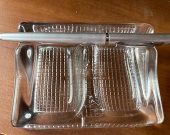 May include: A clear glass pen holder with a grid pattern and a silver pen resting inside. The pen has a silver body and a dark brown tip. The pen holder is rectangular with rounded corners.
