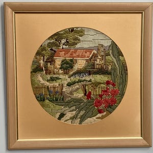 May include: A square gold-toned frame containing a circular needlepoint embroidery depicting a rustic cottage with a red roof, surrounded by green trees, a stone path, and vibrant red flowers in the foreground, all set against a neutral background.