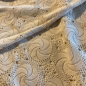 May include: A cream-coloured crocheted textile featuring a repeating spiral pattern. The openwork design creates a delicate, textured surface. The fabric is folded, showcasing the intricate details of the handmade craft. This textile could be used as a tablecloth or decorative element.