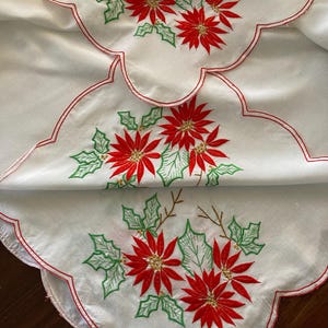 May include: A white fabric runner featuring three embroidered poinsettia clusters with red petals, green leaves, and gold centres. The edges are scalloped and finished with a thin red embroidered border, creating a decorative holiday table accent.