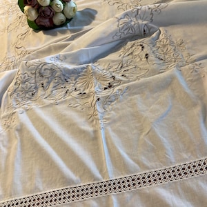 May include: White embroidered tablecloth with floral and leaf patterns. The tablecloth features cutwork and a decorative border. A bouquet of white and burgundy flowers rests on the cloth.