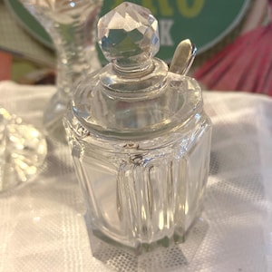 May include: A clear glass condiment jar with a faceted geometric body and a matching lid topped with a pointed, multi-faceted crystal-style finial. The jar features vertical panels and includes a small silver-toned metal spoon tucked inside.