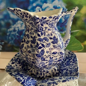 May include: A blue and white floral patterned jug and matching square dish. The jug has a curved handle and a flared spout. The dish is square with a slightly raised edge. The set is likely ceramic.