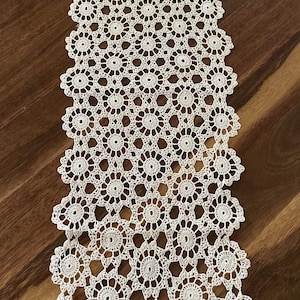 May include: A long, rectangular table runner crafted from off-white thread in a repeating crochet pattern. The design consists of interconnected circular floral motifs with scalloped edges, creating an openwork lace structure throughout the piece.