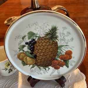 May include: A white ceramic plate with a gold rim, featuring a colourful illustration of a pineapple, grapes, cherries, and other fruits. The plate is displayed on a wooden stand, showcasing a vintage fruit design.