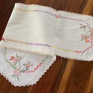 May include: A two-piece set of white embroidered table linens. The rectangular runner and square doily feature floral embroidery in pink, red, yellow, and green. Both pieces have a crocheted lace edge.