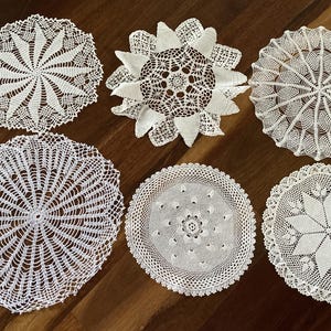 May include: Six white, intricately crocheted doilies in various circular designs are displayed on a dark wood surface. The doilies feature detailed patterns, including starbursts, floral motifs, and geometric shapes, showcasing delicate craftsmanship.