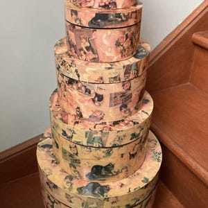 May include: A set of five circular decorative boxes, stacked on top of each other. Each box is adorned with a collage of vintage imagery, featuring children, cats, and floral designs. The boxes vary in size, with the smallest at the top and the largest at the base.
