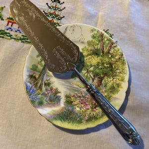 May include: Silver cake server with intricate floral and scroll patterns. The server is placed on a decorative plate depicting a landscape with trees, flowers, and a stream. The plate has a scalloped edge.