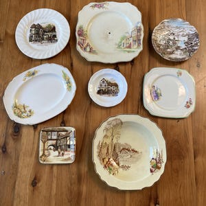 May include: A collection of eight ceramic plates and dishes featuring various shapes and sizes. The set includes round, square, oval, and scalloped pieces decorated with colourful illustrations of buildings, landscapes, and floral patterns.