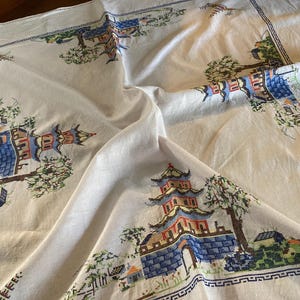 May include: White tablecloth with a detailed cross-stitch pattern featuring pagodas, trees, and architectural elements in shades of blue, red, and green. The border has a geometric design.