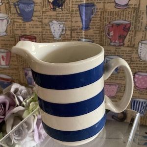 May include: A white ceramic creamer with blue horizontal stripes. It has a handle and a spout. The background features a wallpaper pattern of various teacups.