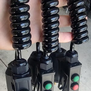May include: Three black industrial pendant control stations, each featuring a green start button and a red stop button. The units are connected to black coiled cables, providing a flexible reach for operating machinery or equipment.