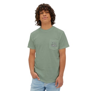 May include: Sage green pocket t-shirt with the text "#1 Best Dad" in black script on the pocket. The shirt is a solid color and has short sleeves. The model is wearing blue jeans.
